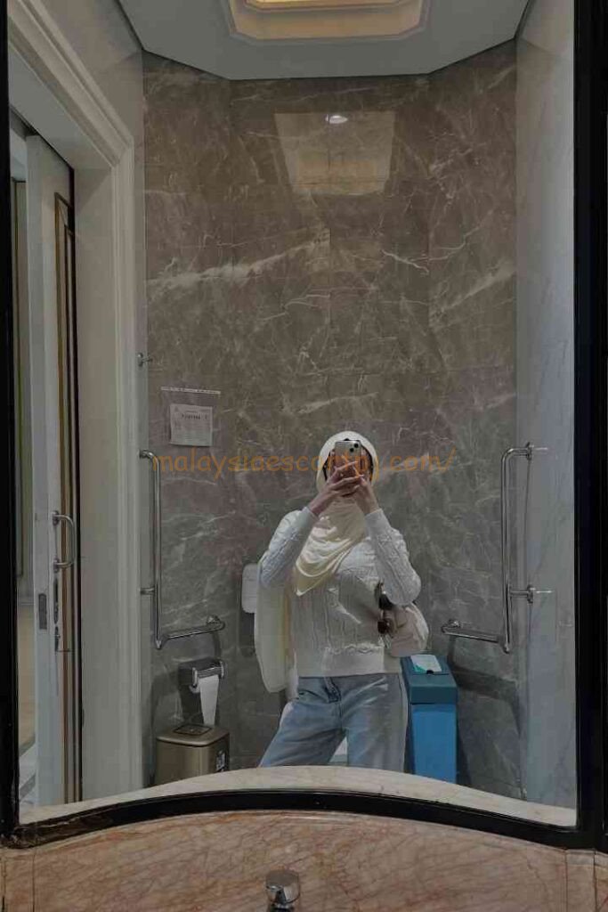 Woman in hijab and white sweater taking a mirror selfie in a modern bathroom